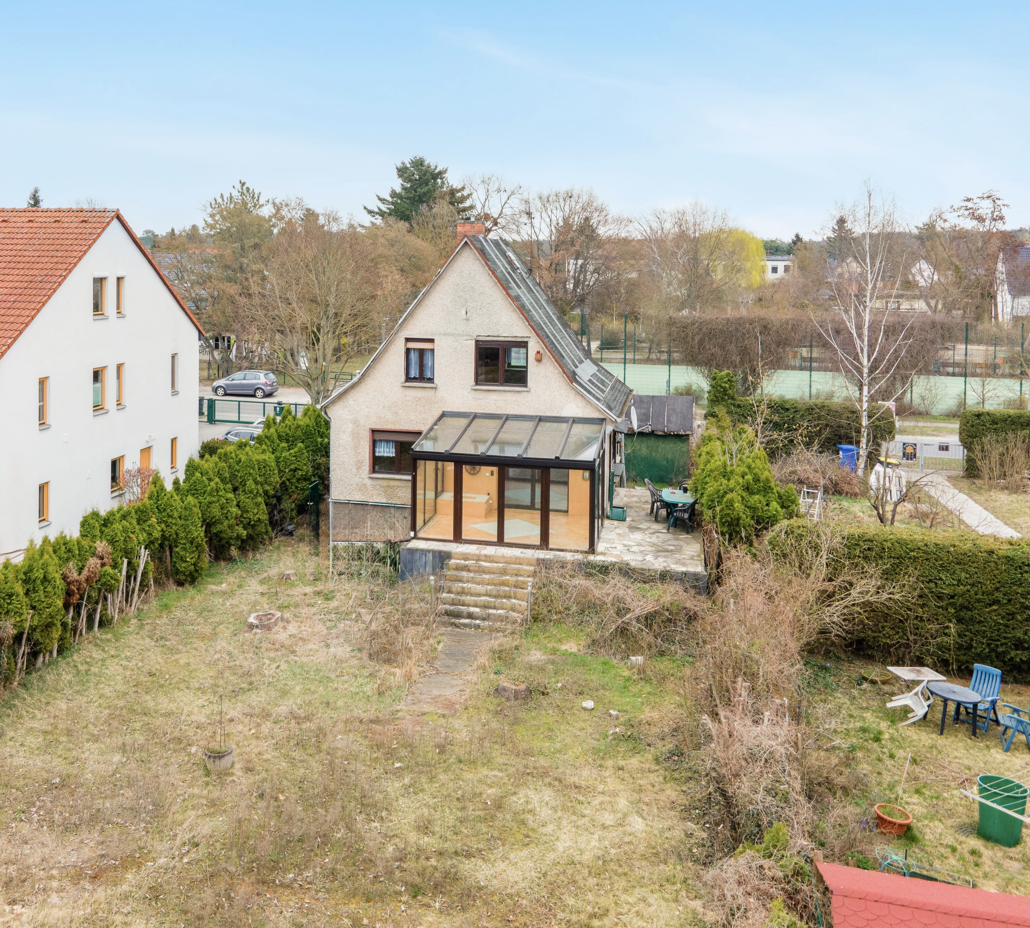 Vorher-Bild für virtuelles Außen-Staging: Ein älteres, renovierungsbedürftiges Einfamilienhaus mit angebautem Glas-Wintergarten und einer einfachen Steinterrasse. Der große, weitläufige Garten wirkt ungepflegt mit kahlem, unebenem Rasen und bietet viel Potenzial für eine digitale Gartengestaltung, Landschaftsarchitektur und Immobilienaufwertung.